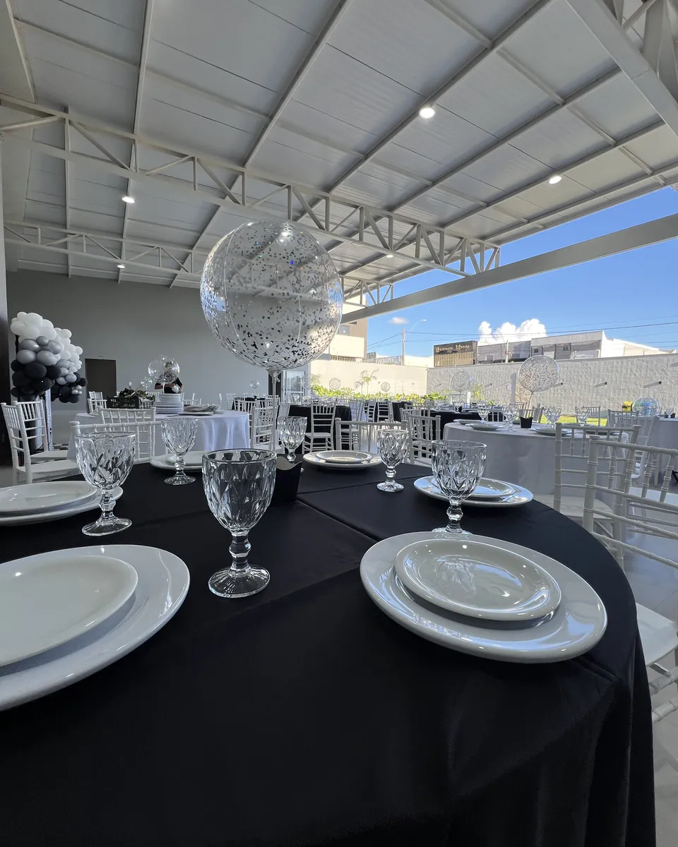 Mesa posta com toalha preta, louça branca e arco de balões ao fundo no salão da Piccolini Eventos