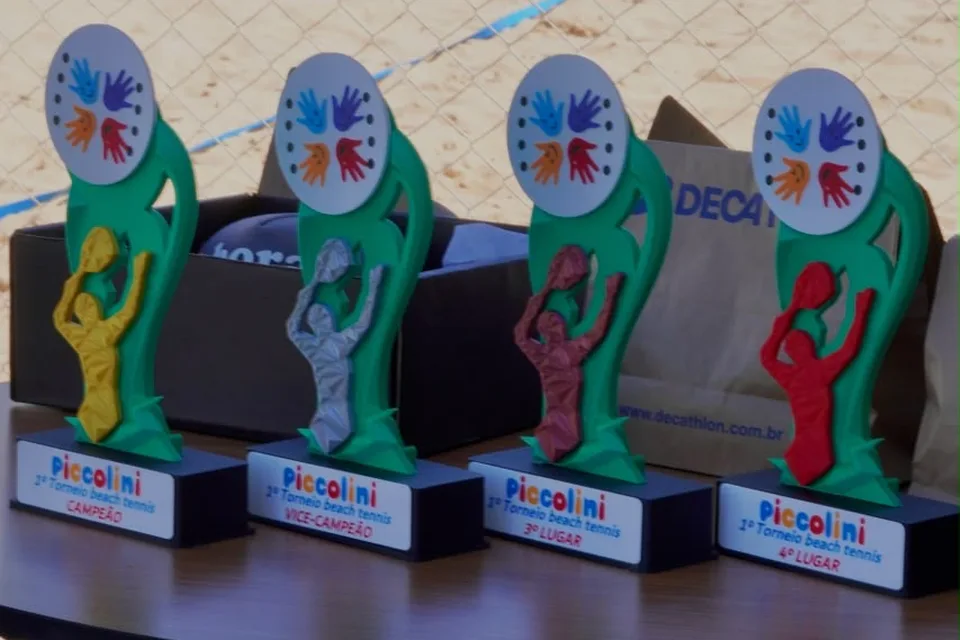 Troféus do 1º Torneio de beach tennis da Piccolini Eventos sobre a mesa, com a quadra de areia ao fundo
